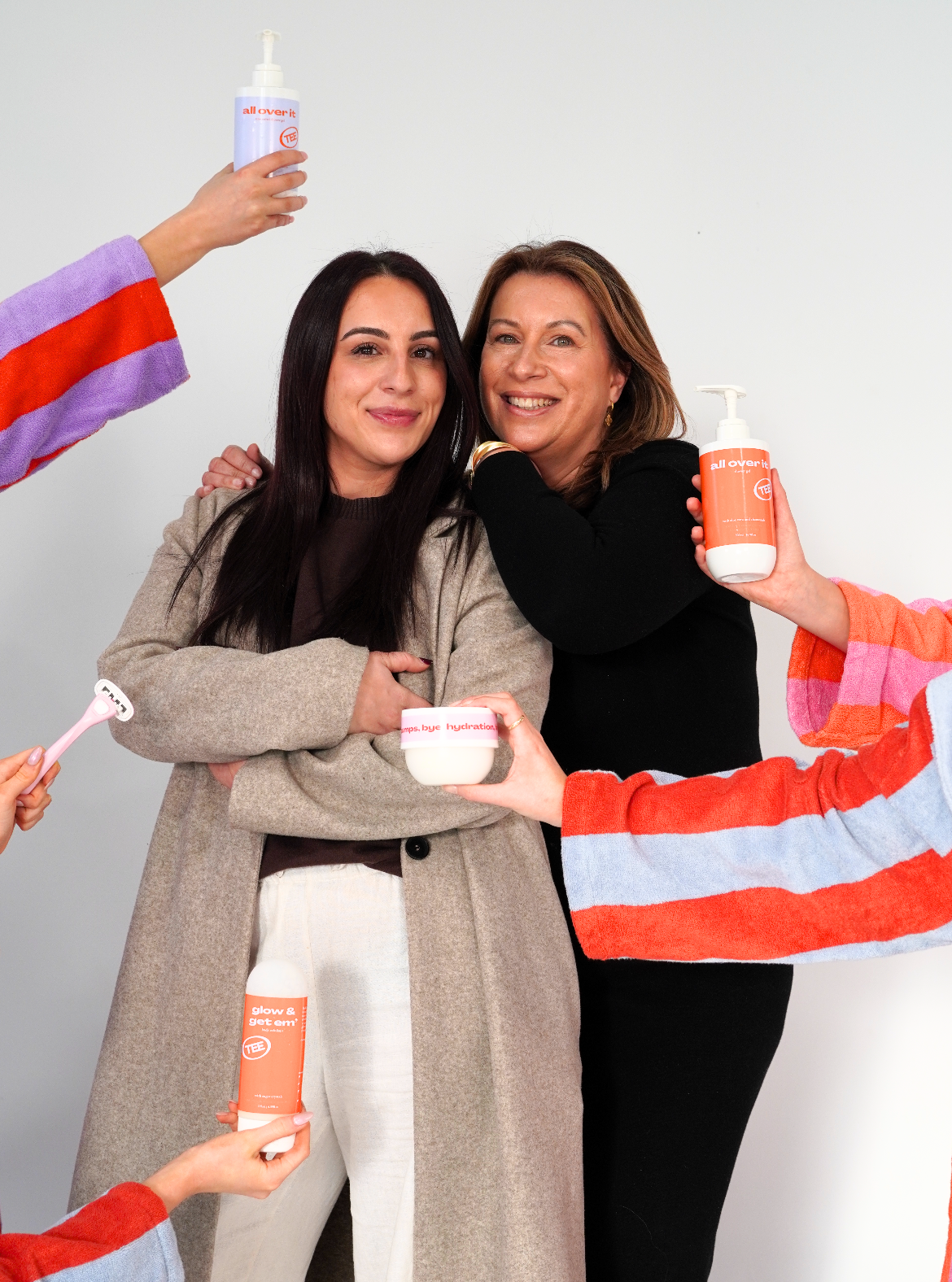 Two women standing together with various products held by people around them on a plain background